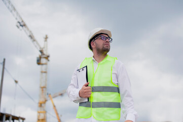 Chief in a vest and a construction helmet with documents in his hands, against the background of a tower crane and the sky with clouds. House construction inspection.