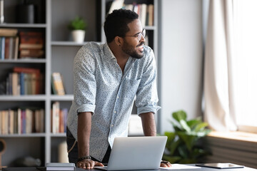 Smiling dreamy African American businessman standing near work desk, looking at window, pensive...