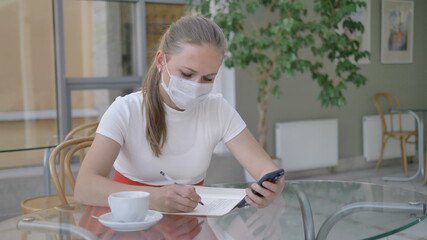 blonde businesswoman with paper notebook and mobile phone takes off medical mask to drink coffee at glass table in cozy cafe
