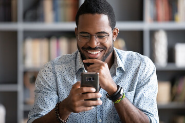 Smiling African American man wearing glasses looking at phone screen, reading good news in message,...