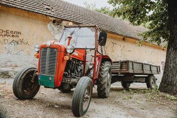 One old red tractor with trailer standing alone in front of building on the gravel street in shadow under the tree during the summer sunny day in Serbia