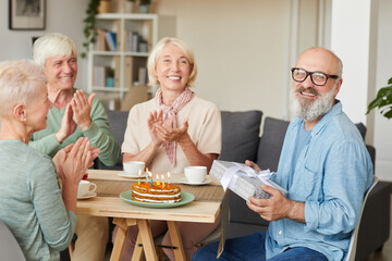 Portrait of senior bearded man holding gift box and smiling at camera while his friends congratulating him at the table