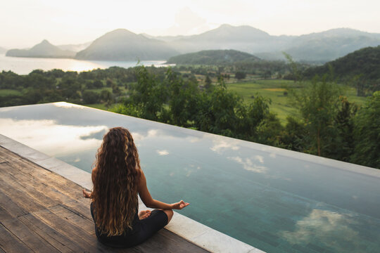 Woman meditating and practicing yoga alone at sunrise near infinity pool with mountains on horizon. Rear view. Travel Lifestyle spiritual relaxation concept. Harmony with nature.