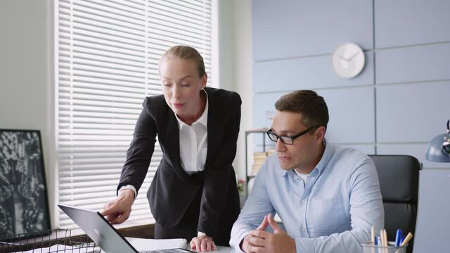female chief stands and male manager sits at table with laptop in office. woman yells and reprimands, looks aggressive and angry, threatens. man is sad and worried.
