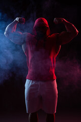 Powerful muscular man with a hood posing in the red sweatshirt. Studio shoot with red and blue smoke
