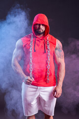 Powerful muscular man with a hood and chain posing in the red sweatshirt. Studio shoot with red and blue smoke