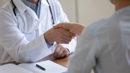 Close up mature doctor wearing white uniform sitting at desk, shaking female patient hand at meeting, senior physician greeting woman at medical appointment, making health insurance agreement