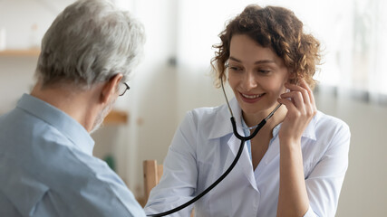Fototapeta premium Smiling young woman doctor using stethoscope close up, listening to mature patient heartbeat or lungs sounds, friendly physician gp consulting senior man at meeting in hospital, medical checkup