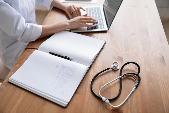 Close Up Young Woman Doctor Working On Laptop In Hospital, Sitting At Table, Female Therapist Physician Gp Work Desk With Stethoscope And Medical Documents, Patient Card, Illness History