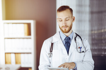 Friendly red-bearded doctor using tablet computer in sunny clinic at his working place. Medicine concept