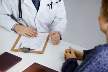 Friendly red-bearded doctor and patient woman discussing current health examination while sitting in sunny clinic. Medicine concept