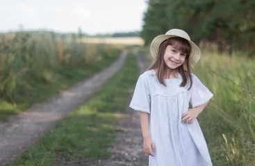Fotobehang Gelukkig meisje op de weg. Schattig meisje in een witte jurk staat in de buurt van het veld © Albert Ziganshin