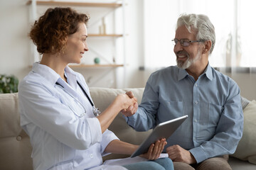 Smiling mature patient shaking doctor nurse hand, thanking, greeting at meeting, homecare concept, young woman caregiver congratulating senior man with good medical checkup result, using tablet