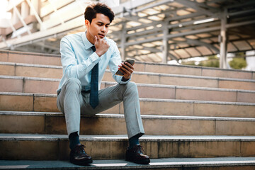 Young Striving Asian Businessman Using Mobile Phone in the City. Sitting on Staircase. Reading Seriously topic via Smartphone
