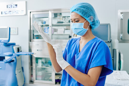 Vietnamese Female Surgeon In Medical Mask Putting On Rubber Gloves And Getting Ready For Surgery