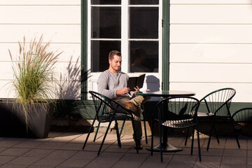 
Full length horizontal shot of young man sitting at a restaurant terrace reading during a late summer sunny afternoon