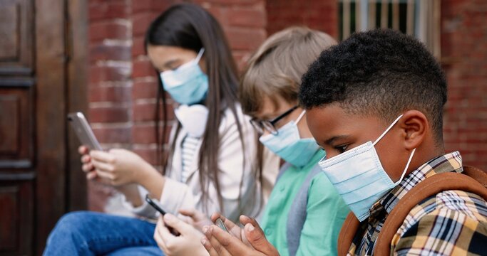 Close up portrait of multi-ethnic children in masks with backpacks standing outdoor and tapping on smartphones.Caucasian girl and African American boy browsing and playing on cellphones School concept