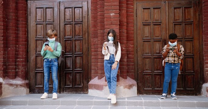 Multi-ethnic Children In Masks With Backpacks Standing Outdoor Near School And Tapping On Smartphones. Caucasian Girl And Boy Browsing On Cellphone. African American Kid Playing On Device Kids Concept