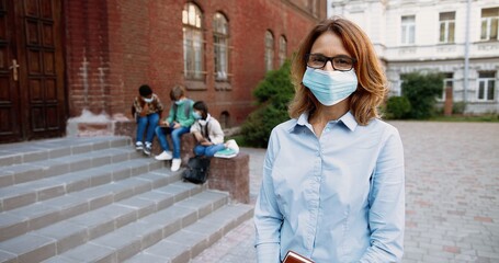Portrait of cheerful beautiful Caucasian female teacher in mask smiling to camera in front of...