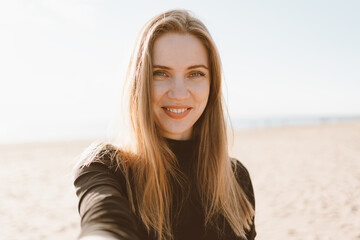 Pretty female with long hair, blonde takes photo on mobile phone on sandy beach in summer or autumn. Beautiful woman looking at camera and smiling in sunny day in ocean or sea coastline