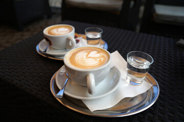Two cups of cappuccino coffee with artistic latte art and glass of tap water in luxury modern cafe, where 100% bio arabica is used