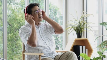 Happy senior male sitting on chair wear red headphone for listen the music and sing a song together in living room, feeling fun and happy. Activity lifestyle after retirement concept.