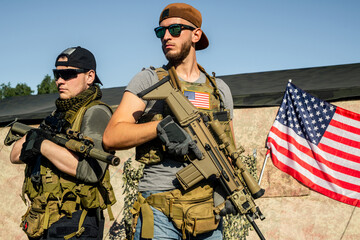 Purposeful young men in sunglasses standing with modern rifles at shooting range, American flag on...