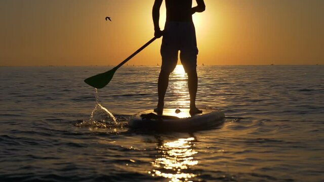 man standing firmly on SUP board and paddling, pushing the paddle through shining water and propelling paddle board at sunrise