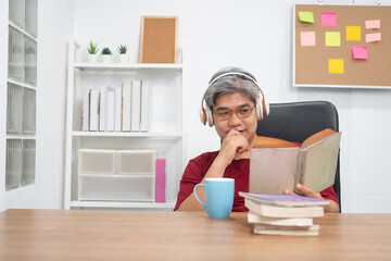 Relax elderly Asian man reading a book and listening to music in home office.