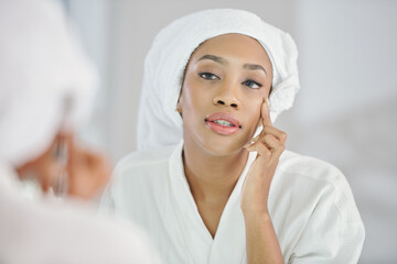Beautiful young Black woman looking at mirror and checking undereye wrinkles after morning shower