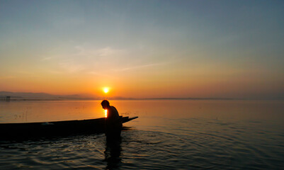 At lake side, asian fisherman sitting on boat and using fishing rod to catch fish at the sunrise