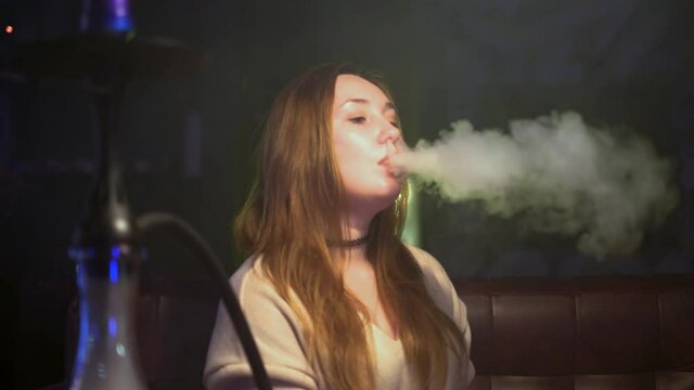 Beautiful woman exhales smoke from hookah. Media. Woman enjoys smoking hookah and slowly exhales smoke. Woman smokes beautifully in hookah bar