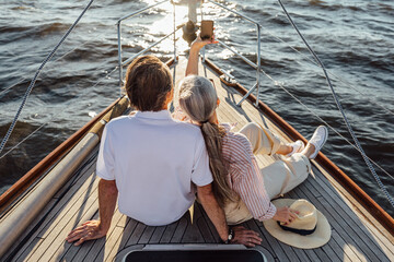 Rear view of senior man and woman sitting on private yacht and taking selfie with mobile phone