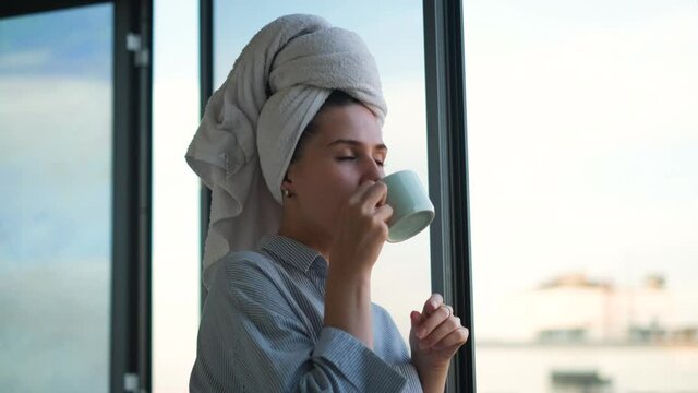 Woman with towel on head drinking coffee in background of window. Concept. Beautiful young woman preparing for start of working day with cup of coffee on balcony