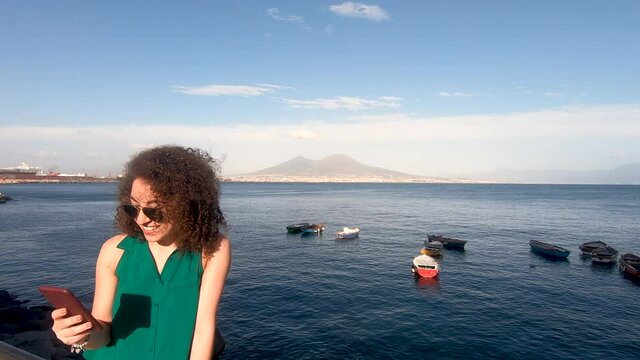 Pretty young curly woman, stands near naples sea during clear cloudy summer evening. She is hipster and trendy with sunglass, chats on smartphone. Vesuvio vulcano and some boat in background, Napoli.