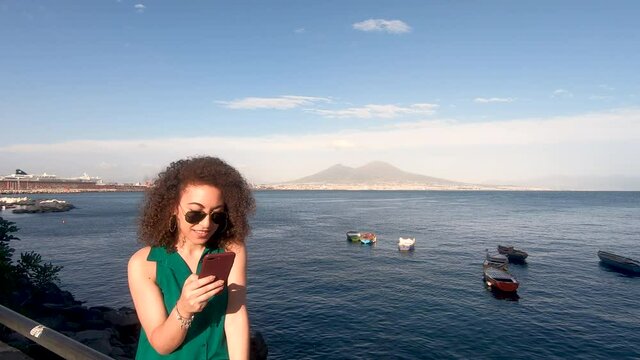 Pretty young curly hair woman, stands near naples sea during clear cloudy summer evening. She is hipster and trendy with sunglass, chats on smartphone. Vesuvio vulcano and some boat in background, Nap