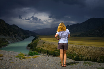 Young woman in white t-shirt standing back to the camera in the sunlight and looks at the beautiful view of Katun river, plains and rocks. In the distance, torrents of rain, overcast sky.