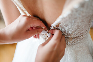 Bridesmaid helping bride fasten buttons on corset and getting her dress, preparing bride in morning for the wedding day.