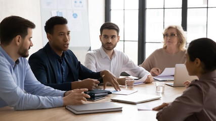 Focused mixed race business people of different ages listening to african american colleague, talking at meeting. Young biracial male specialist explaining marketing strategy to teammates in office.