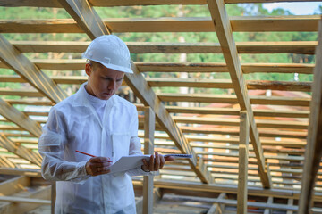 Fototapeta premium Foreman holding drawings, looking the project of wooden house, working on a construction site. Young attractive engineer in a white helmet checks the roof frame. Concept career architect. Copy space.