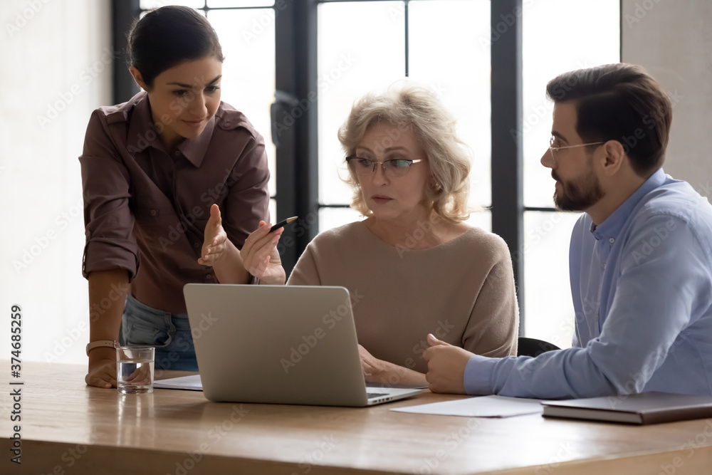 Wall mural Focused middle aged 60s businesswoman discussing online project presentation with multiracial young colleagues at brainstorming meeting. Diverse employees developing company growth strategy in office.