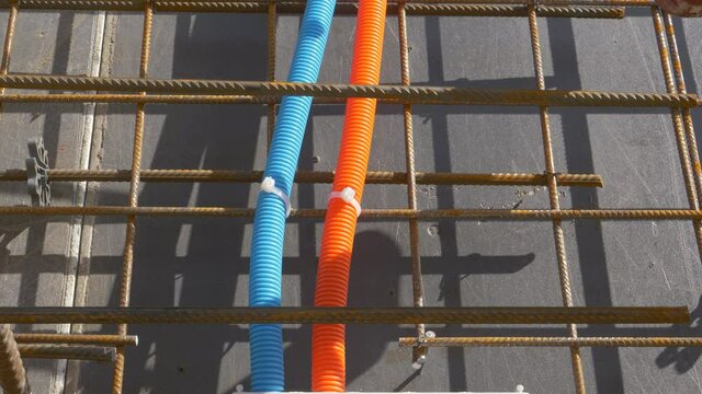 CLOSE UP: Two colorful electricity and internet conduits run behind the rusty metal wiring covering the black insulation sheet covering an unfinished housing project. Two corrugated plastic tubes.