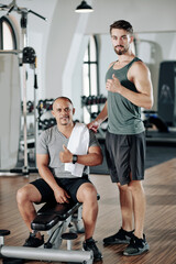 Handsome young fitness trainer and his client showing thumbs-up and looking at camera after finishing working out in gym