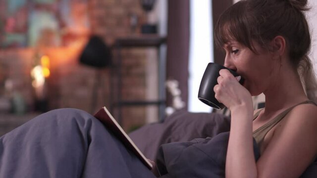 Woman at home lying in bed at morning drinking coffee and reading book. High quality 4K video footage.