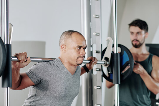 Serious fitness trainer controlling his mature client doing squats with heavy barbell