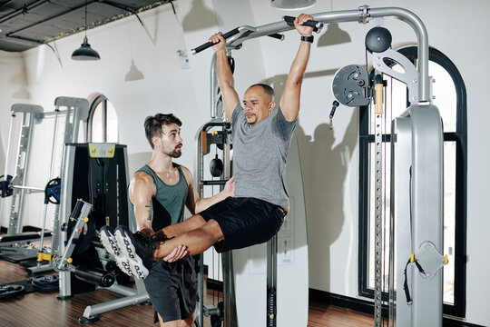 Personal sports trainer controlling client raising straight legs when hanging on gym bar