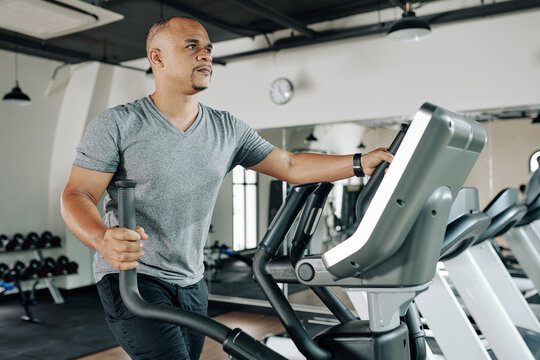 Serious mature mixed-race man working out on elliptical machine in gym to warm up before training