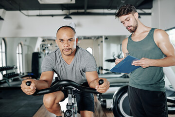 Fototapeta na wymiar Mature mixed-race man riding fast on stationary bicycle in gym, his personal trainer taking notes in document