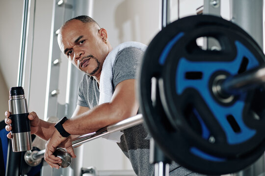 Frowning serious strong man leaning on barbell, resting and drinking water between sets - Powered by Adobe