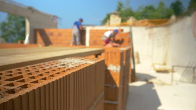 CLOSE UP, DOF: Blurry shot of contractors laying bricks and building walls behind a short brick wall in the middle of the first floor of a house under construction. Bricklayers at work on a sunny day.